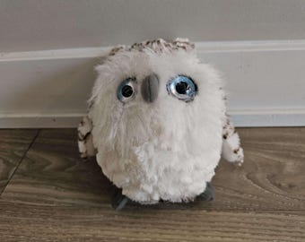 May include: A white plush owl with blue eyes and brown accents. The owl is sitting on a wooden floor.