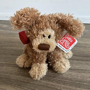 May include: A plush brown dog with a red bow and a tag that reads "Gotta Getta GUND". The stuffed animal has a shaggy appearance and is sitting on a wooden surface.