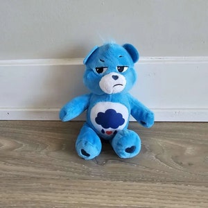 May include: Blue Care Bear plush toy with a sad expression and a dark blue cloud with a raindrop on its chest.