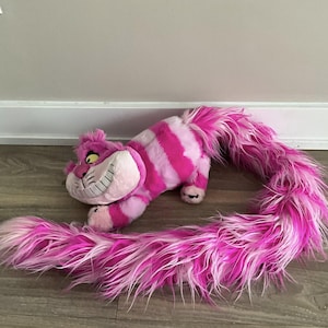 May include: A plush Cheshire Cat toy from Alice in Wonderland. The cat is pink and white striped with a long, fluffy tail. It has a large, smiling face with yellow eyes and a wide grin. The toy is lying on a wooden floor.