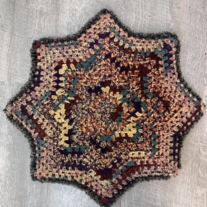 May include: A star-shaped crochet mat featuring a multi-colored yarn pattern in shades of deep burgundy, muted teal, soft yellow, and earthy brown. The edges are finished with a dark, fuzzy, textured yarn border that outlines the six-pointed shape.