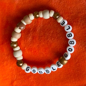 May include: A beaded bracelet with a mix of wooden and gold-colored beads. The bracelet spells out the word "GOBBLE" using white letter beads. The beads are strung together on an elastic band, creating a festive accessory.