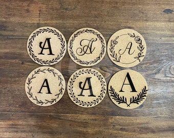 May include: Six round cork coasters with a black floral wreath design and the letter "A" in the center. The coasters are on a wooden surface.