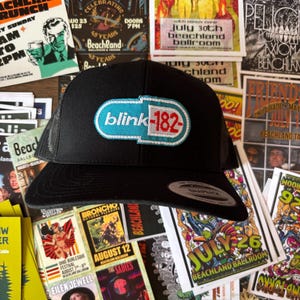 May include: Black trucker hat featuring the band name "blink-182" in a teal and red design. The hat is surrounded by various concert posters and flyers, creating a music-themed aesthetic.