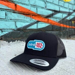 May include: Black snapback hat with a mesh back and a flat brim. The hat features a white, light blue, and red logo that says "blink-182". The hat has a snap closure and a silver sticker on the brim.