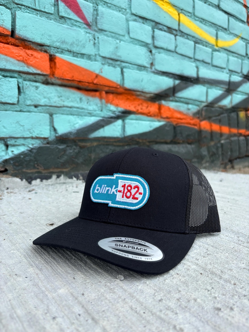 May include: Black snapback hat with a mesh back and a flat brim. The hat features a white, light blue, and red logo that says "blink-182". The hat has a snap closure and a silver sticker on the brim.