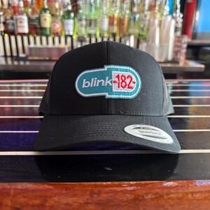 May include: Black snapback hat featuring the band name "blink-182" in a teal and red design. The hat has a curved brim and a mesh back. The hat is sitting on a wooden surface.
