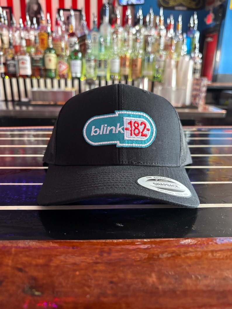 May include: Black snapback hat featuring the band name "blink-182" in a teal and red design. The hat has a curved brim and a mesh back. The hat is sitting on a wooden surface.