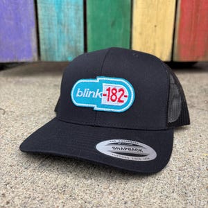 May include: Black snapback hat with a mesh back and a curved brim. The hat features a white, light blue, and red logo that says "blink-182". The hat has a snap closure and a Yupoong tag.