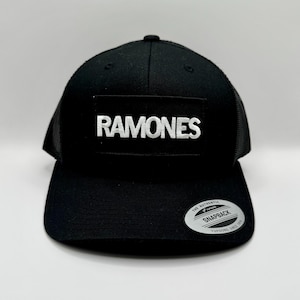 Könnte beinhalten: Schwarze Snapback-Kappe mit Mesh-Rückseite und flachem Schirm. Auf der Vorderseite befindet sich ein rechteckiger Aufnäher mit dem weißen Wort "RAMONES". Ein silberner und schwarzer "Snapback"-Aufkleber befindet sich auf dem Schirm.