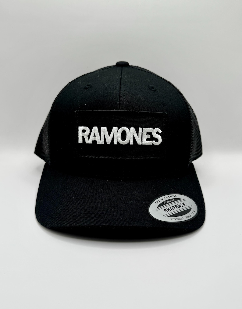K&ouml;nnte beinhalten: Schwarze Snapback-Kappe mit Mesh-R&uuml;ckseite und flachem Schirm. Auf der Vorderseite befindet sich ein rechteckiger Aufn&auml;her mit dem wei&szlig;en Wort "RAMONES". Ein silberner und schwarzer "Snapback"-Aufkleber befindet sich auf dem Schirm.