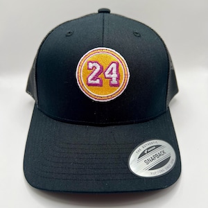 May include: Black mesh trucker hat with a yellow and pink embroidered patch featuring the number "24". The hat has a black brim and a black adjustable strap with a silver "The Authentic Yupoong Snapback Yupoong Since 1974" tag.