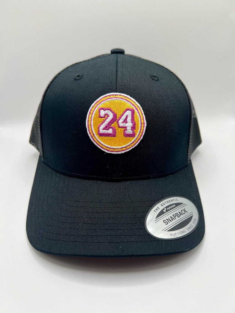 May include: Black mesh trucker hat with a yellow and pink embroidered patch featuring the number "24". The hat has a black brim and a black adjustable strap with a silver "The Authentic Yupoong Snapback Yupoong Since 1974" tag.