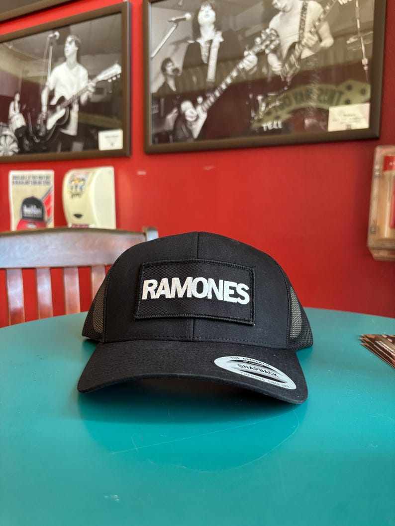 K&ouml;nnte beinhalten: Schwarze Trucker-M&uuml;tze mit dem Wort "RAMONES" in wei&szlig;en Blockbuchstaben auf einem rechteckigen Aufn&auml;her. Die M&uuml;tze hat eine Netzr&uuml;ckseite und einen gebogenen Schirm. Die M&uuml;tze liegt auf einer t&uuml;rkisfarbenen Oberfl&auml;che.
