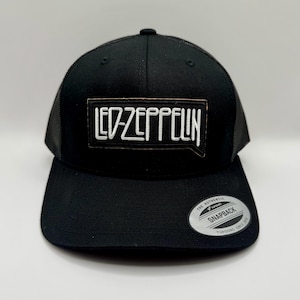 Puede incluir: Gorra de camionero negra con visera curva y parte trasera de malla. La parte delantera presenta un parche rectangular con el nombre de la banda "LED-ZEPPELIN" en letras blancas. Una pegatina plateada y negra "SNAPBACK" está en la visera.