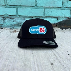 May include: Black trucker hat with a curved brim and mesh back. The front features a white, blue, and red logo that reads "blink-182". The hat is sitting on a concrete surface.