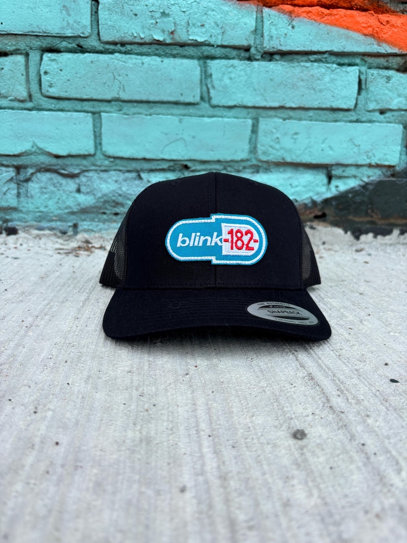 May include: Black trucker hat with a curved brim and mesh back. The front features a white, blue, and red logo that reads "blink-182". The hat is sitting on a concrete surface.