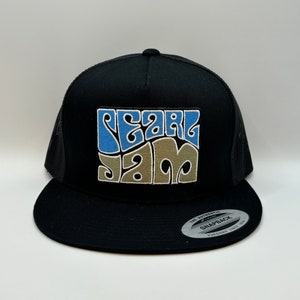 May include: Black trucker hat with a black mesh back. The front of the hat features a white and blue embroidered patch that says "Pearl Jam" in a psychedelic font.