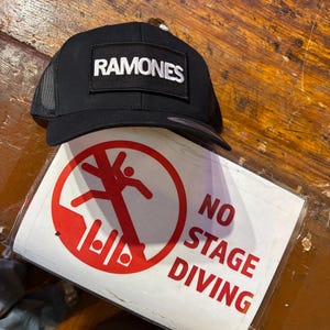 K&ouml;nnte beinhalten: Schwarze Trucker-Cap mit dem Wort "RAMONES" in wei&szlig;en Buchstaben. Ein Schild mit einem roten Kreis und einer Strichm&auml;nnchen-Figur sowie den Worten "NO STAGE DIVING" ist ebenfalls zu sehen.