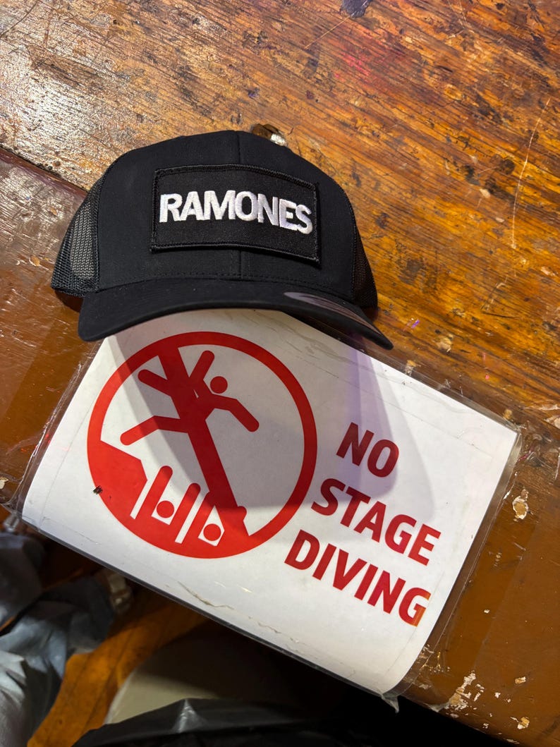 K&ouml;nnte beinhalten: Schwarze Trucker-Cap mit dem Wort "RAMONES" in wei&szlig;en Buchstaben. Ein Schild mit einem roten Kreis und einer Strichm&auml;nnchen-Figur sowie den Worten "NO STAGE DIVING" ist ebenfalls zu sehen.