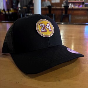 May include: Black trucker hat with a yellow and purple patch that says "24" on it.