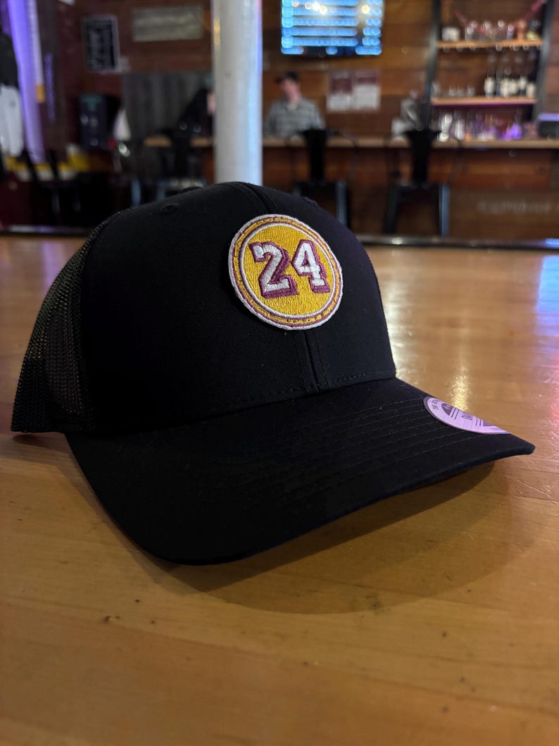 May include: Black trucker hat with a yellow and purple patch that says "24" on it.