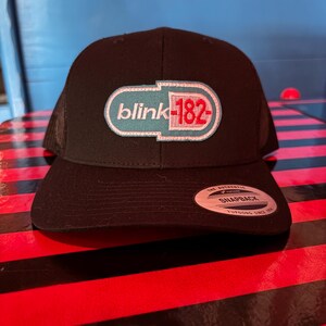 May include: Black snapback hat with a mesh back and a curved brim. The front features a white, teal, and red embroidered logo that reads "blink-182". A white tag with black text is on the brim.