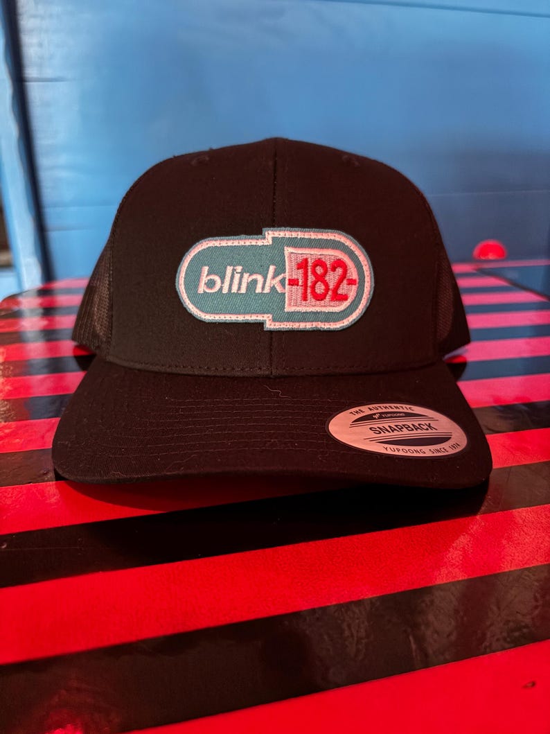 May include: Black snapback hat with a mesh back and a curved brim. The front features a white, teal, and red embroidered logo that reads "blink-182". A white tag with black text is on the brim.