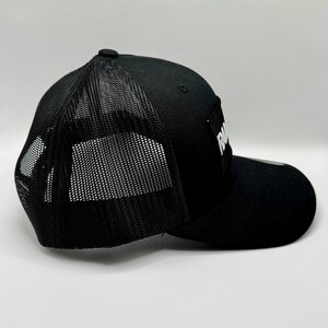 K&ouml;nnte beinhalten: Schwarze Trucker-Kappe mit Mesh-R&uuml;ckseite und gebogenem Schirm. Die Kappe hat einen wei&szlig;en rechteckigen Aufn&auml;her mit dem Wort "RA" in wei&szlig;en Blockbuchstaben. Mit verstellbarem Snapback-Verschluss.