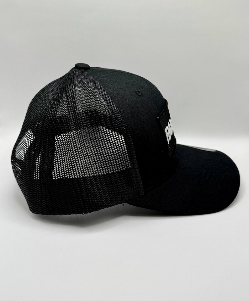 K&ouml;nnte beinhalten: Schwarze Trucker-Kappe mit Mesh-R&uuml;ckseite und gebogenem Schirm. Die Kappe hat einen wei&szlig;en rechteckigen Aufn&auml;her mit dem Wort "RA" in wei&szlig;en Blockbuchstaben. Mit verstellbarem Snapback-Verschluss.