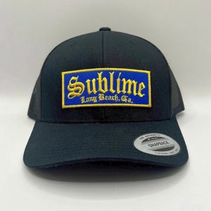 May include: Black trucker hat with a mesh back and a curved brim. The front features a rectangular patch with the word "Sublime" in yellow script over a blue background, with "Long Beach, Ca." below.