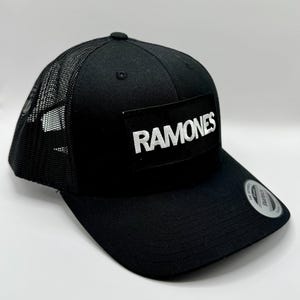 K&ouml;nnte beinhalten: Schwarze Trucker-Kappe mit Mesh-R&uuml;ckseite und gebogenem Schirm. Auf der Vorderseite der Kappe befindet sich ein rechteckiger Aufn&auml;her mit dem Wort "RAMONES" in wei&szlig;en Blockbuchstaben. Ein kleines rundes Etikett befindet sich auf dem Schirm.