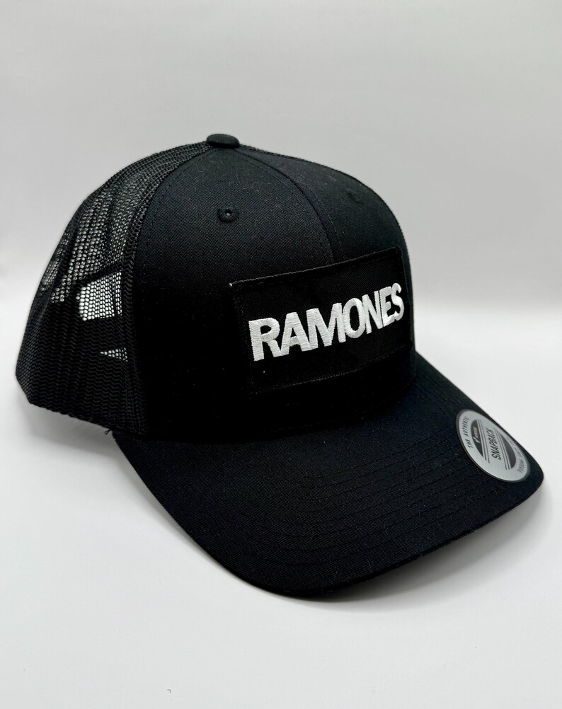 K&ouml;nnte beinhalten: Schwarze Trucker-Kappe mit Mesh-R&uuml;ckseite und gebogenem Schirm. Auf der Vorderseite der Kappe befindet sich ein rechteckiger Aufn&auml;her mit dem Wort "RAMONES" in wei&szlig;en Blockbuchstaben. Ein kleines rundes Etikett befindet sich auf dem Schirm.