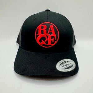 Może przedstawiać: Czarna czapka typu trucker z czerwonym haftowanym logo "RAGE" w okrągłym wzorze. Czapka posiada zakrzywiony daszek, siatkowy tył i regulowane zapięcie na zatrzask. Biała metka z czarnym tekstem znajduje się na daszku.