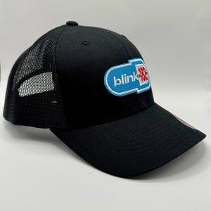 May include: Black trucker hat with a mesh back and a curved brim. The front features a patch with the text "blink-182" in white, blue, and red. The hat is a classic baseball cap style.