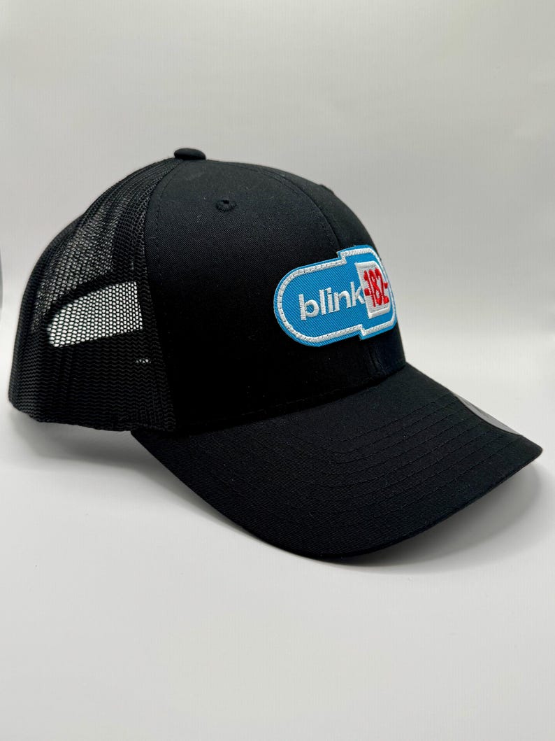 May include: Black trucker hat with a mesh back and a curved brim. The front features a patch with the text "blink-182" in white, blue, and red. The hat is a classic baseball cap style.