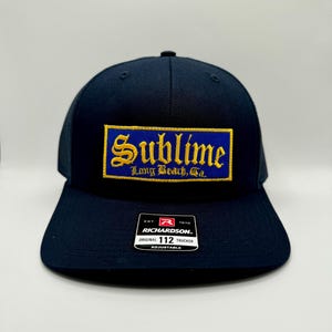 Op de afbeelding: Marineblauwe truckerpet met een mesh achterkant en een gebogen klep. De voorkant heeft een rechthoekige patch met het woord "Sublime" in gouden letters op een blauwe achtergrond. De tekst "Long Beach, Ca." staat eronder. Een Richardson 112-label staat op de klep.