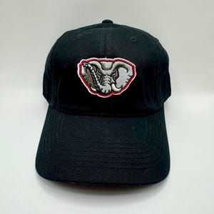 May include: Black baseball cap with a curved brim. The cap features an embroidered patch of an elephant head in gray and white, outlined in red, centered on the front. The cap is made of a durable fabric.