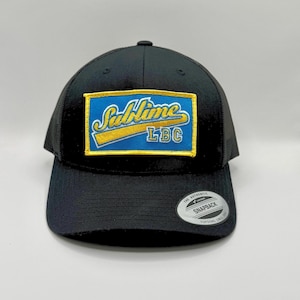 May include: Black trucker hat with a mesh back and a curved brim. The front features a rectangular patch with the word "Sublime" in yellow script over a blue background, with "LBC" below. A "Snapback" sticker is on the brim.