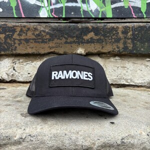 K&ouml;nnte beinhalten: Schwarze Trucker-M&uuml;tze mit Mesh-R&uuml;ckseite und gebogenem Schirm. Die M&uuml;tze hat einen rechteckigen Aufn&auml;her mit dem Wort "RAMONES" in wei&szlig;en Blockbuchstaben. Die M&uuml;tze liegt auf einer Steinoberfl&auml;che.