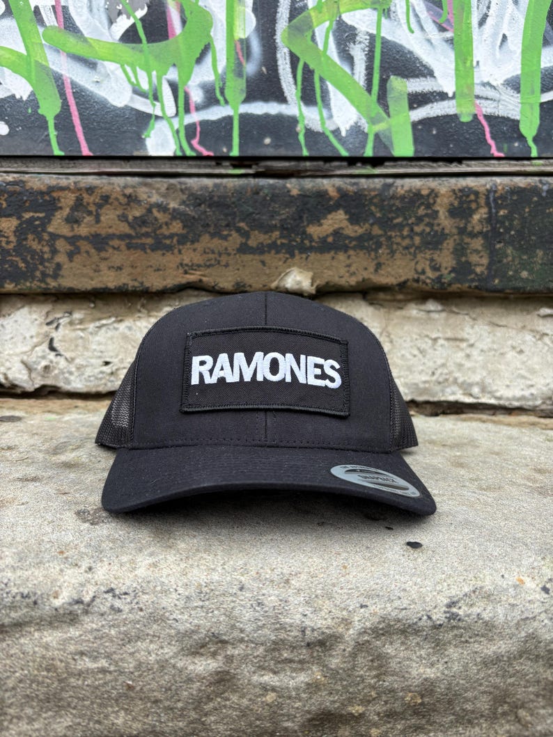 K&ouml;nnte beinhalten: Schwarze Trucker-M&uuml;tze mit Mesh-R&uuml;ckseite und gebogenem Schirm. Die M&uuml;tze hat einen rechteckigen Aufn&auml;her mit dem Wort "RAMONES" in wei&szlig;en Blockbuchstaben. Die M&uuml;tze liegt auf einer Steinoberfl&auml;che.