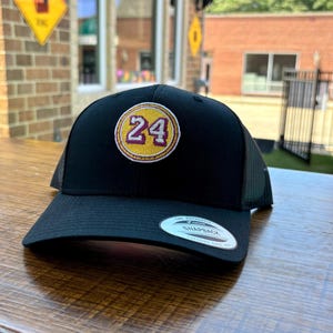 May include: Black mesh trucker hat with a yellow and burgundy embroidered patch featuring the number 24.