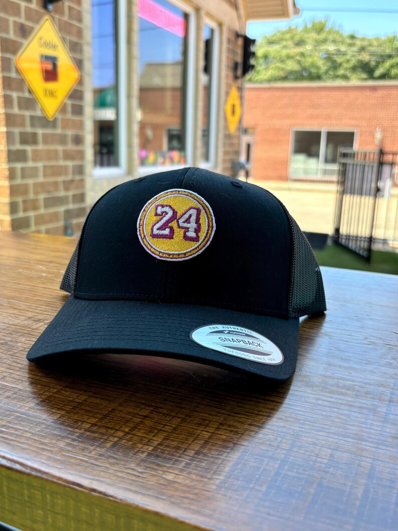 May include: Black mesh trucker hat with a yellow and burgundy embroidered patch featuring the number 24.
