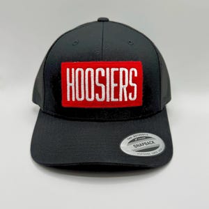 May include: Black trucker hat with a red rectangular patch featuring the word "HOOSIERS" in white block letters. The hat has a curved brim and a mesh back. A silver "SNAPBACK" sticker is on the brim.