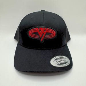 May include: Black snapback hat with a red embroidered logo. The hat features a curved brim and a mesh back. The logo is an oval shape with a stylized "VF" design. The hat has a "Yupoong" tag.