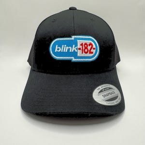 Könnte beinhalten: Schwarze Snapback-Kappe mit dem Bandnamen "blink-182" in Weiß, Rot und Blau. Die Kappe hat einen gebogenen Schirm und eine Mesh-Rückseite. Auf dem Schirm befindet sich ein silberner Aufkleber.