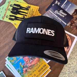 K&ouml;nnte beinhalten: Schwarze Trucker-Cap mit Mesh-R&uuml;ckseite und flachem Schirm. Die Kappe hat einen rechteckigen Aufn&auml;her mit dem Wort "RAMONES" in wei&szlig;en Blockbuchstaben. Die Kappe liegt auf einer Holzoberfl&auml;che mit Konzertflyern im Hintergrund.