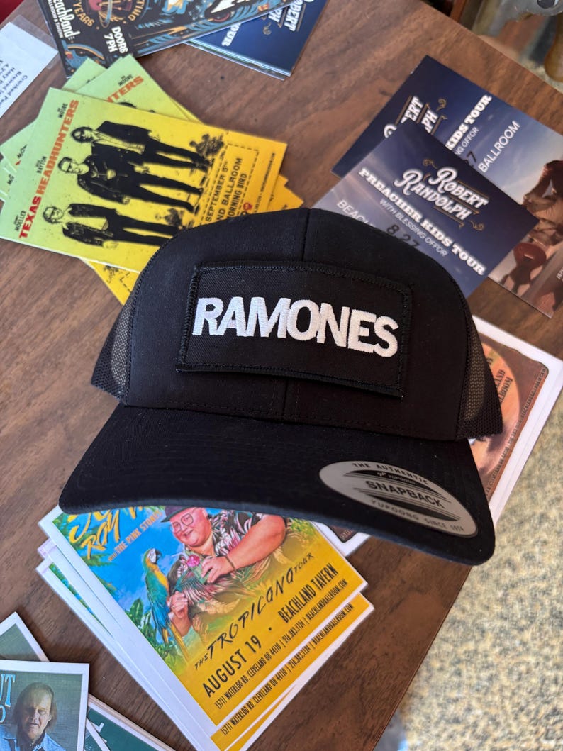 K&ouml;nnte beinhalten: Schwarze Trucker-Cap mit Mesh-R&uuml;ckseite und flachem Schirm. Die Kappe hat einen rechteckigen Aufn&auml;her mit dem Wort "RAMONES" in wei&szlig;en Blockbuchstaben. Die Kappe liegt auf einer Holzoberfl&auml;che mit Konzertflyern im Hintergrund.