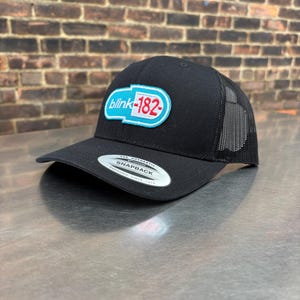 May include: Black snapback hat with a mesh back and a curved brim. The hat features a patch with the text "blink-182" in white and red on a blue oval. The hat is made by Yupoong.