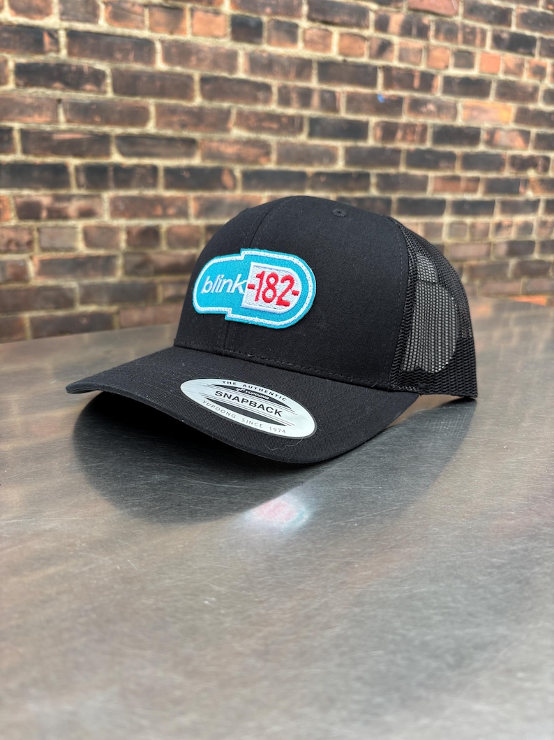 May include: Black snapback hat with a mesh back and a curved brim. The hat features a patch with the text "blink-182" in white and red on a blue oval. The hat is made by Yupoong.